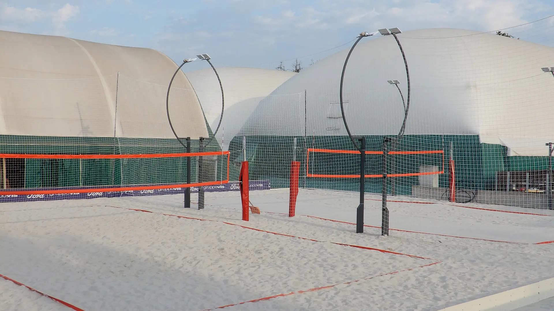 Campi da beach volley con sabbia chiara, reti e linee arancioni e illuminazione su pali a profilo curvo, in un centro sportivo